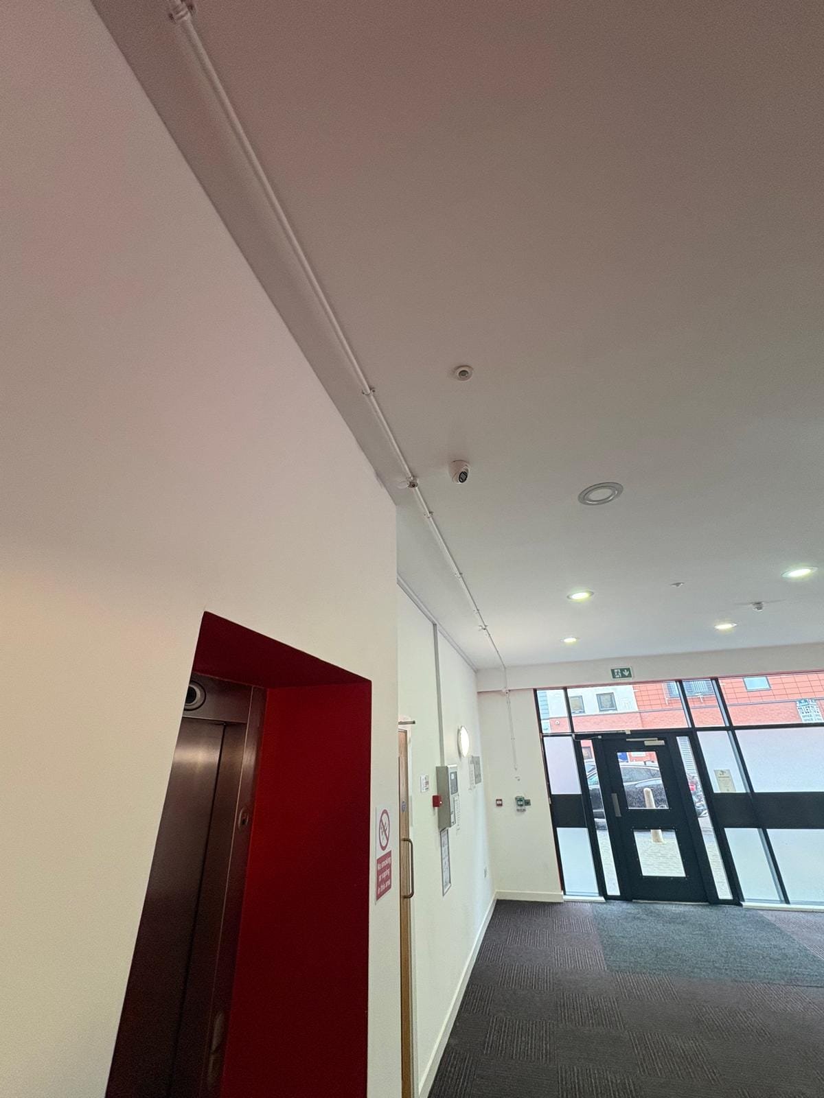 Ground floor corridor with surface-mounted conduit runs and ceiling CCTV near the entrance doors