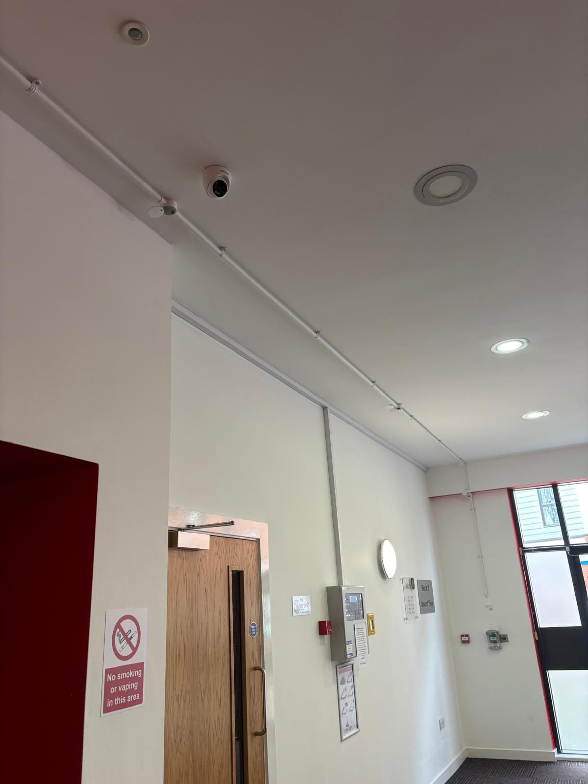 Communal corridor with surface conduit, ceiling CCTV dome and fire alarm panel routed along the wall